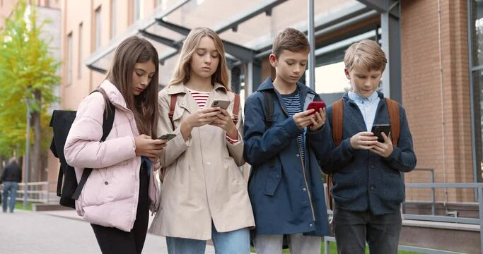 Caucasian concentrated teens standing outdoors and typing on cellphones. Beautiful girl pupil browsing on smartphone near school. Young boy school student texting on mobile phone. Friends concept