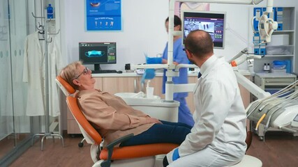 Assistant preparing patient for dental intervention putting dental bib while doctor specialist talking with senior woman pointing on digital screen explaining x-ray of teeth. - Powered by Adobe