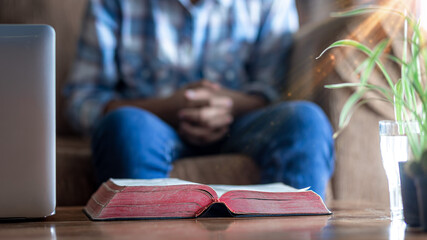 Close up christian Bible on wooden table with young male praying in background, Home church during...