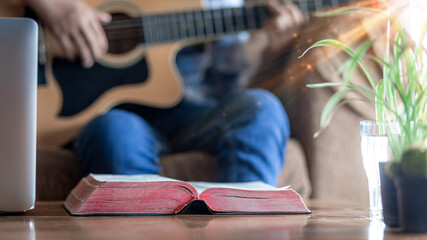 Close up christian Bible on wooden table with young male playing guitar for worship God in background, Home church during quarantine coronavirus Covid-19, Christian concept. - obrazy, fototapety, plakaty