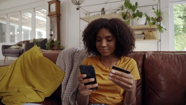 Camera panning over african woman sitting on couch holding cellphone using credit card to make instant payment