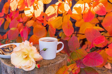 Obraz na płótnie Canvas Cup of coffee, pastries, and roses on a wooden stump among the orange leaves of the scumpia tree in autumn