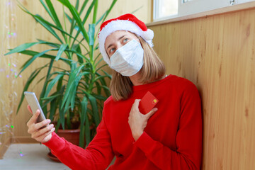 Christmas online shopping: a happy woman in a red Santa hat and medical mask makes an order online and pays with a credit card through the phone. gifts via online stores.