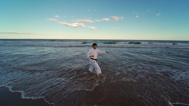 Karate fighter practicing katas on the seashore. Karate fighter practicing techniques with his feet in the sea at sunset. Karate kicking and punching in a powerful way. Traditional karate.