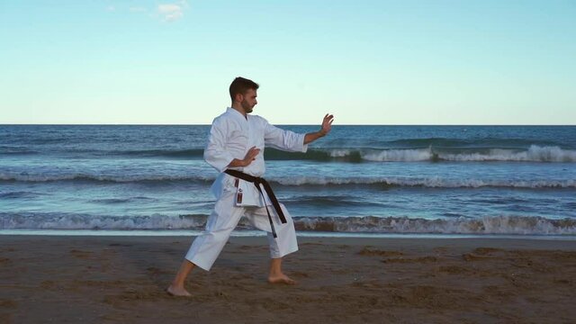 Karate fighter practicing katas on the beach at sunset and doing very energetic and powerful techniques on the seashore. Wado ryu style. Truck right camera movement recorded in slow motion