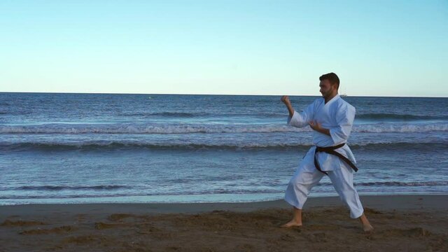 Karate fighter practicing katas on the beach at sunset and doing very powerful techniques and doing a flying kick on the seashore. Wado ryu style. Truck left camera movement recorded in slow motion