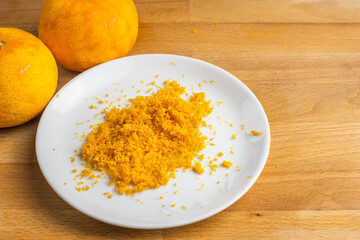 Grated orange zest on a white plate on a wooden tabletop with two oranges without the outer skin.