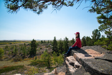 A girl in a red cap sits on a rock above a forest and field.