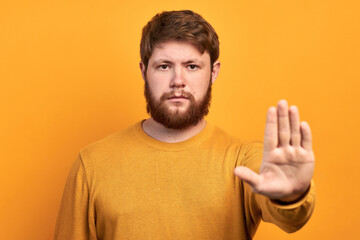 a young bearded man looking serious, angry and displeased forbidding to enter or saying stop, forbidding with his hand on a yellow background
