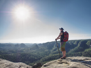 Hiker with red backpack on cliff hold trekking sticks. Cowhoy watching morning landscape against sun