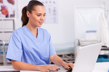 Medical physician typing on laptop sitting at desk wearing blue uniform. Health care physician...