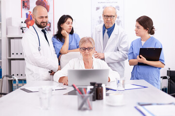 Fototapeta premium Mature doctor specialist briefing her medical team in conference room using laptop. Clinic expert therapist talking with colleagues about disease, medicine professional