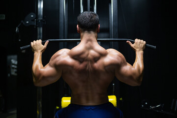 Handsome fitness man is performing back workouts using thrust of the upper block machine in a gym, back view
