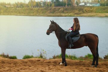 Cute young woman on horseback in autumn forest by lake. Rider female drives her horse in Park in inclement cloudy weather with rain. Concept of outdoor riding, sports and recreation. Copy space