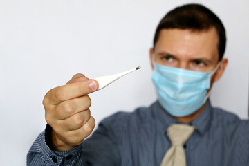Worried man in face mask and office clothes looks at digital thermometer in hand. High body...