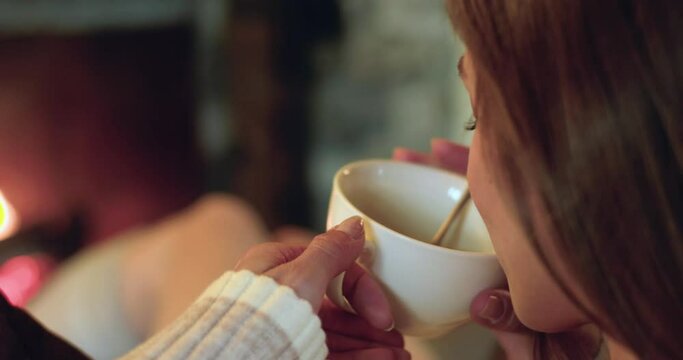 Back view of young woman having coffee while relaxing at home during cold winter. Woman sitting on sofa looking fireplace.