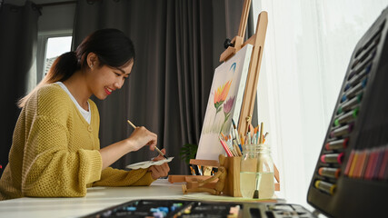 Fototapeta premium Portrait of happy young woman artist holding palette and paintbrush while working on painting in her workshop.