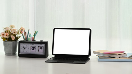 Modern workspace with blank screen tablet, notebooks, clock and tree pot on white table.