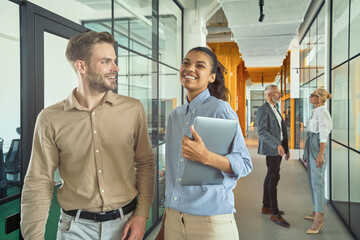 Two young happy multiracial male and female coworkers talking and smiling while walking in the modern office