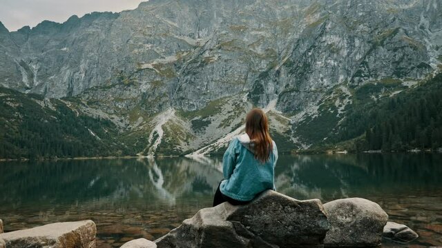 Young tourist woman rest on a mountain lake, enjoys landscape high in the mountains, back view
