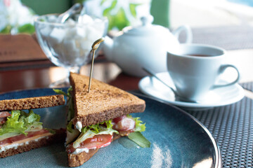 sandwiches with bacon herbs and tomatoes on a dark plate with a white Cup of tea a teapot and a sugar bowl in the background with a blurred background