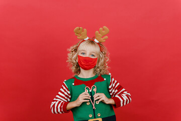 Merry Christmas on quarantine coronavirus pandemic. Little girl with sweet lollipop candy cane in Santa elf helper costume wearing medicine mask on red vivid color background.