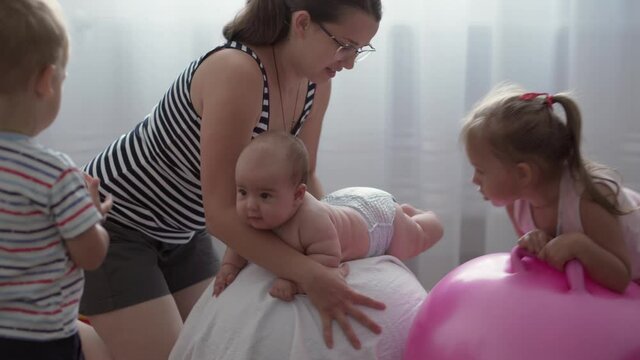 Childhood, Motherhood, games, upbringing and development concept - close-up young mom are joyfully doing exercise and masah with two newborn and little daughters on fitball and smiling in kid playroom