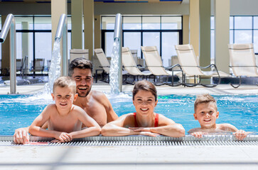 Smiling family of four having fun and relaxing in indoor swimming pool at hotel resort.