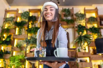Female waitress wearing face shield, visor serves the coffee in restaurant during coronavirus pandemic representing new normal concept