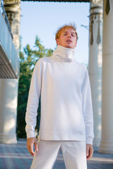 Handsome young man in fashionable white clothes. Stands stylishly against the backdrop of the sky and modern architecture.