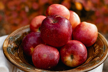 Juicy ripe red apples in autumn against the background of seasonal red colorful leaves. Crunchy sweet fruit in a vintage ceramic plate with a white tablecloth. Delicious fruits of appetizing color