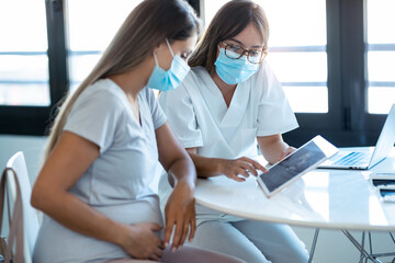 Young beautiful gynecologist wearing a hygienic face mask while showing to pregnant woman ultrasound scan baby with digital tablet in medical consultation.