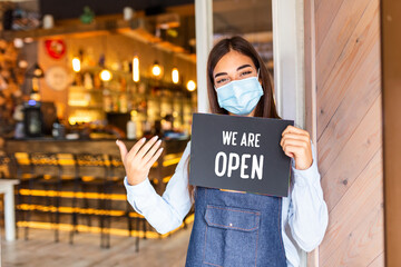 Happy female waitress with protective face mask holding open sign while standing at cafe or restaurant doorway, open again after lock down due to outbreak of coronavirus covid-19