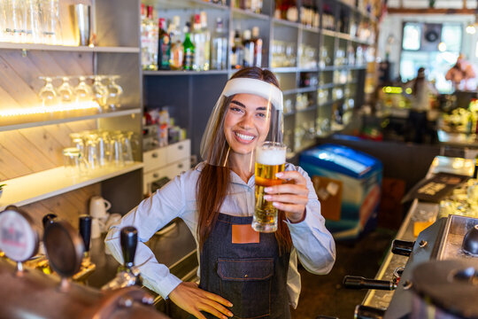 Bartender with face shield,covid-19 protection, serving a draft beer at the bar counter during coronavirus pandemic, showing OK sign,shelves full of bottles with alcohol on the background