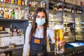 Beautiful female Bartender wearing protective face mask, serving a draft beer during coronavirus pandemic, shelves full of bottles with alcohol on the background