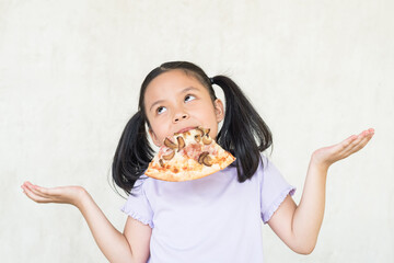 surprised little asia girl keeps slice of pizza in mouth, spreads hands with hesitation, has...