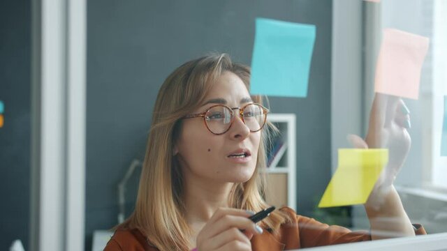 Slow motion of creative young businesswoman writing info on sticky notes on glass board working alone in office. Business planning and marketing concept.