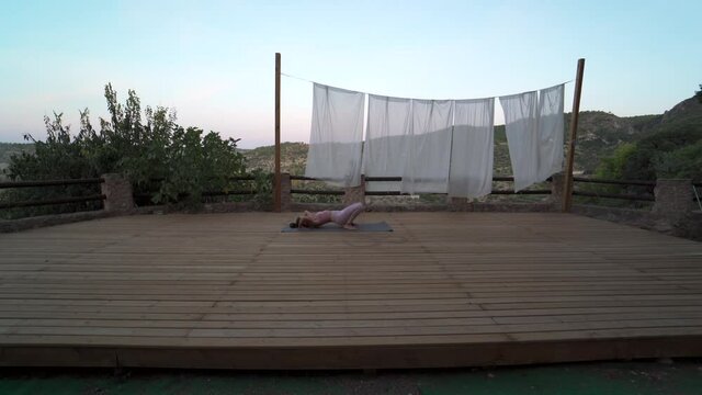 Woman practicing yoga in nature on a wooden platform mountains in the background. Unique natural setting at sunset. Woman practice upward bow pose and one legged bow pose. Eke pada urdhva dhanurasana