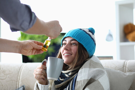 Young woman addict sits in hat and scarf on sofa at home with cup with marijuana extract portrait. Illegal cannabis treatment at home concept.