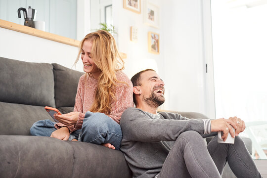 Happy young couple laughing holding cups of coffee and using phone on the couch together in the living room.