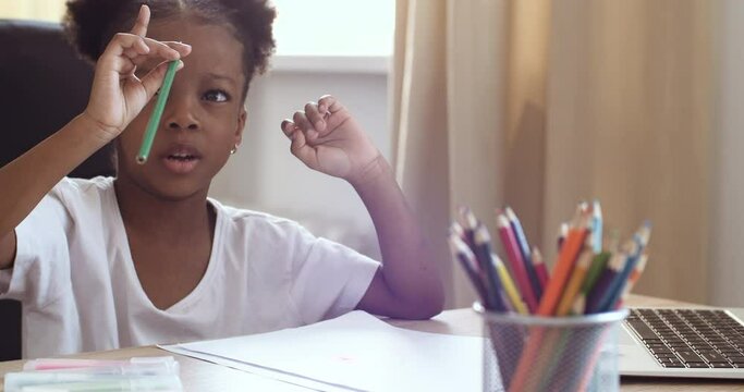 Little cute african girl with curly hair sits at table at home, plays alone, draws on paper, chooses colored pencil, engaged in creativity studies remotely, comments her picture, home learning concept