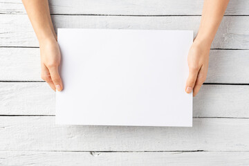 Overhead shot of woman’s hands holding blank paper sheet on white wooden table. Close up