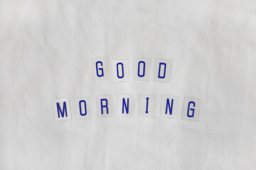Fototapeta na wymiar Inscription Good morning, composed of dark blue letters on transparent plastic bases on white rumpled sheets. Top view, flat lay. Horizontal. Concept of rest, awakening, sleep. Minimal style