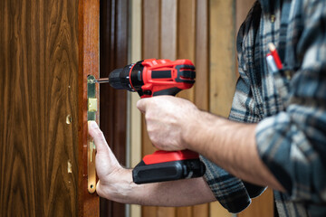 Close-up. Carpenter with an electric screwdriver fixes the lock of a wooden door. Construction...