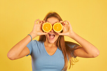 happy young woman using oranges as glasses on a yellow background