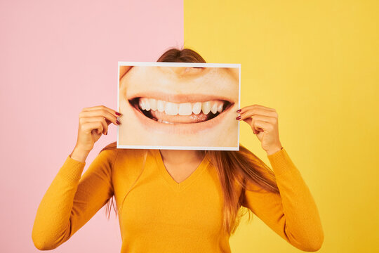 young woman holding a picture of mouth smiling showing her teeth on yellow background. Dentist concept