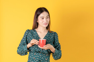 Beautiful young Asian woman hold coffee feel warm with hot red cup stand on yellow background in studio, portrait attractive lady happy with coffee/ tea. lifestyle working woman entrepreneur concept