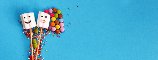 marshmallows on wooden skewers with color sweets on blue background. holiday concept. sweets in form of people. banner