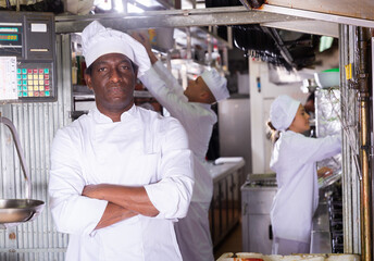 Fototapeta na wymiar Experienced African American chef of restaurant posing with arms crossed in kitchen on background with working staff