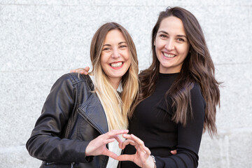 Two smiling girls hugging and forming a heart with their hands. They are out in the open.